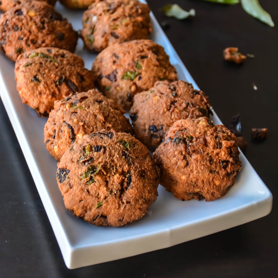 A white rectangular plate filled with golden-brown fried lentil fritters, garnished with green herbs.