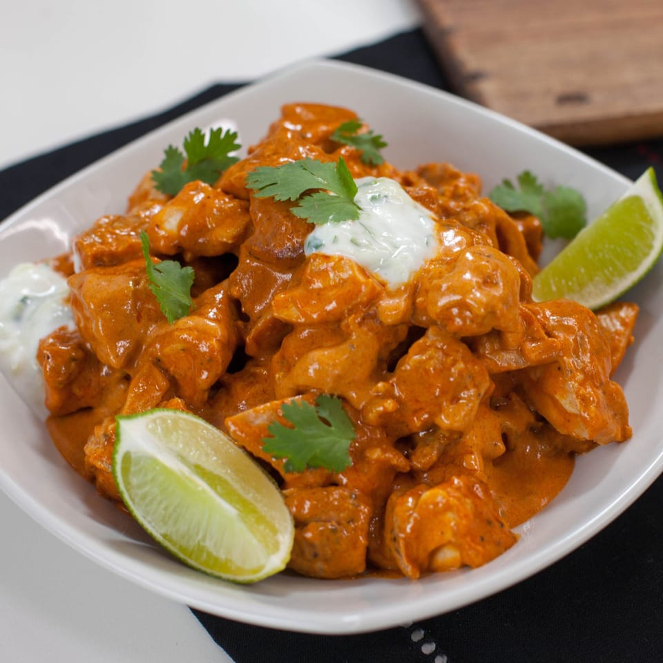 Chicken tikka masala with cilantro, lime wedges, and a dollop of raita in a white bowl.