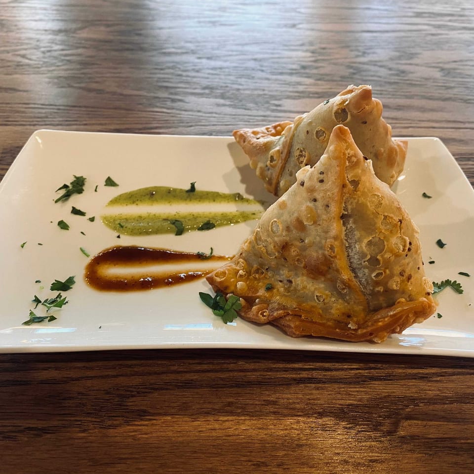 Two golden-brown samosas on a white rectangular plate with green and brown sauces, garnished with parsley.